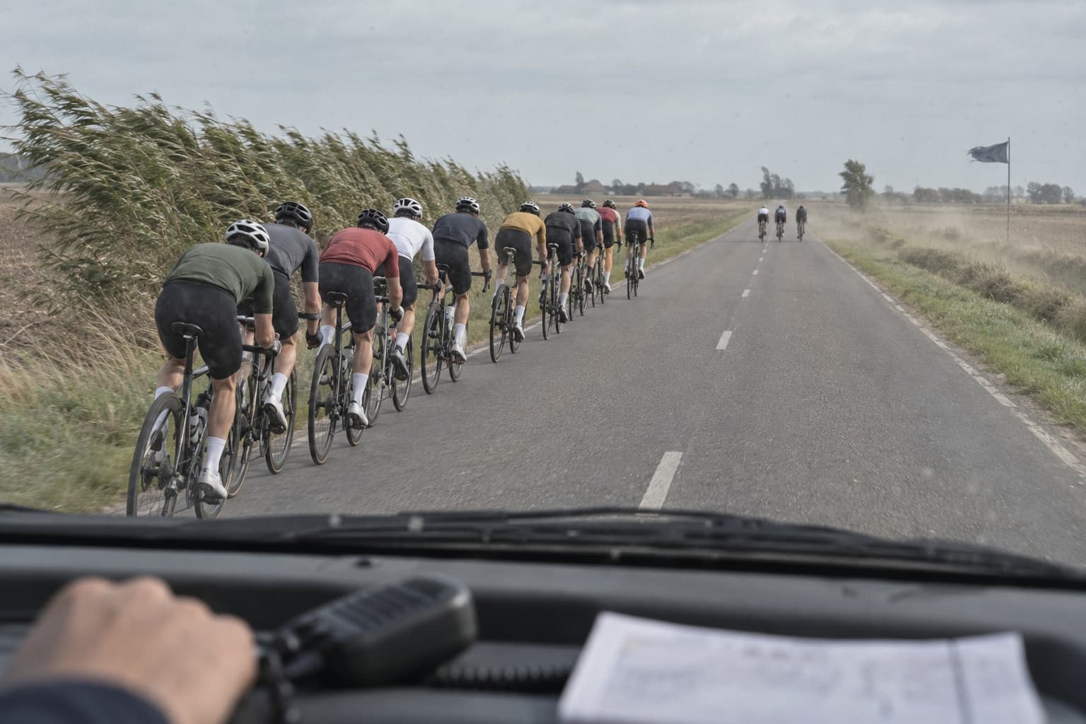 Uma longa fila de ciclistas numa estrada plana e exposta, vegetação dobrada pelo vento