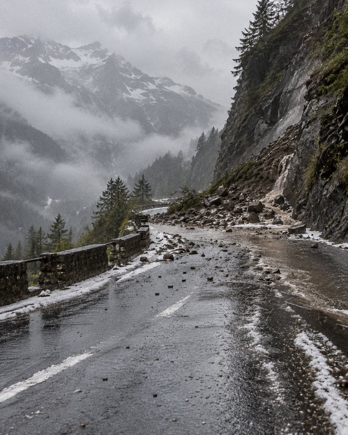 A mountain road half-buried by a fresh landslide, rock and mud across the tarmac under low cloud and hail